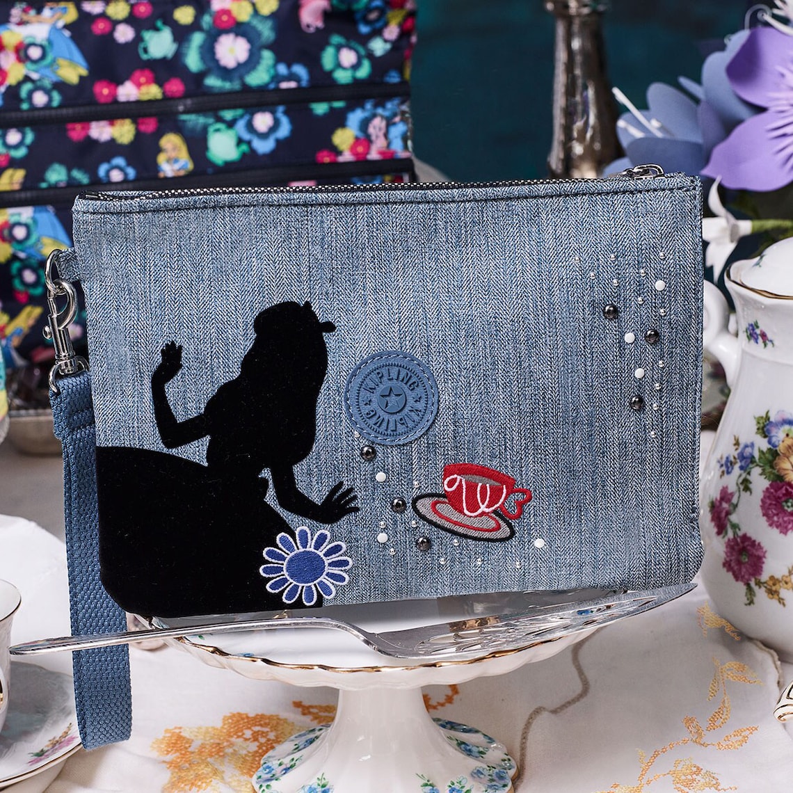 A Kipling denim wristlet pouch featuring a silhouette of Alice in Wonderland and an embroidery flower and tea cup on display among a tea serving set