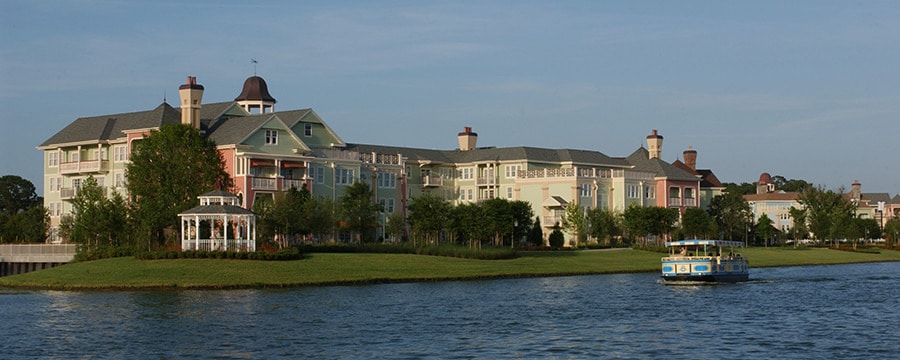 An open aired riverboat sailing alongside the Victorian style Disney's Saratoga Springs Resort and Spa