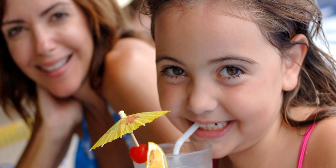 A girl sips an umbrella topped tropical drink with her mom in the background A girl sips an umbrella topped tropical drink with her mom in the background