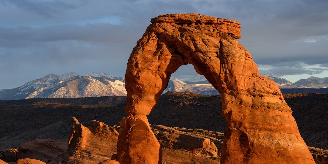 A natural sandstone arch in Arches National Park with snow-capped mountains in the background A natural sandstone arch in Arches National Park with snow-capped mountains in the background