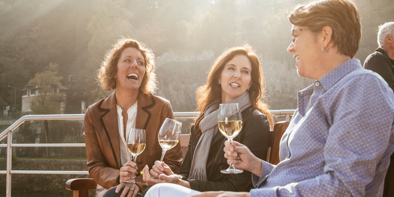 Three women with glasses of wine enjoy a discussion while seated on an outside deck of a river boat as they pass a tree-lined shore