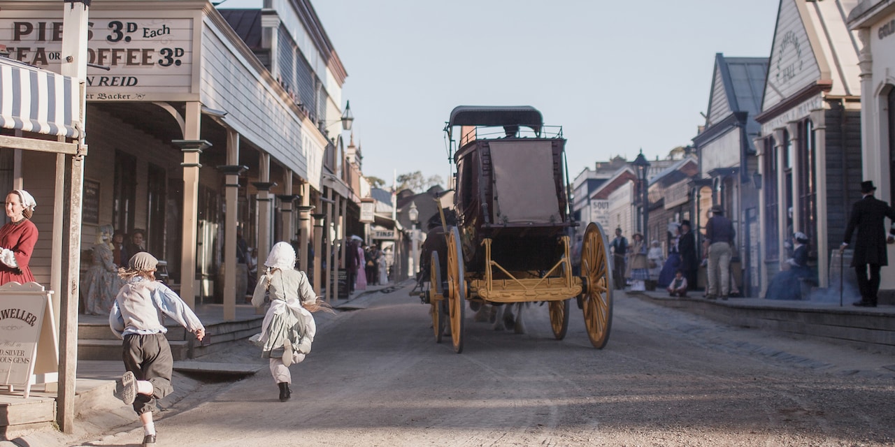 Two kids run after a horse drawn buggy traveling down a dirt road lined with shops and people dressed in 1800s era clothing at Sovereign Hill Open Air Museum in Melbourne, Australia