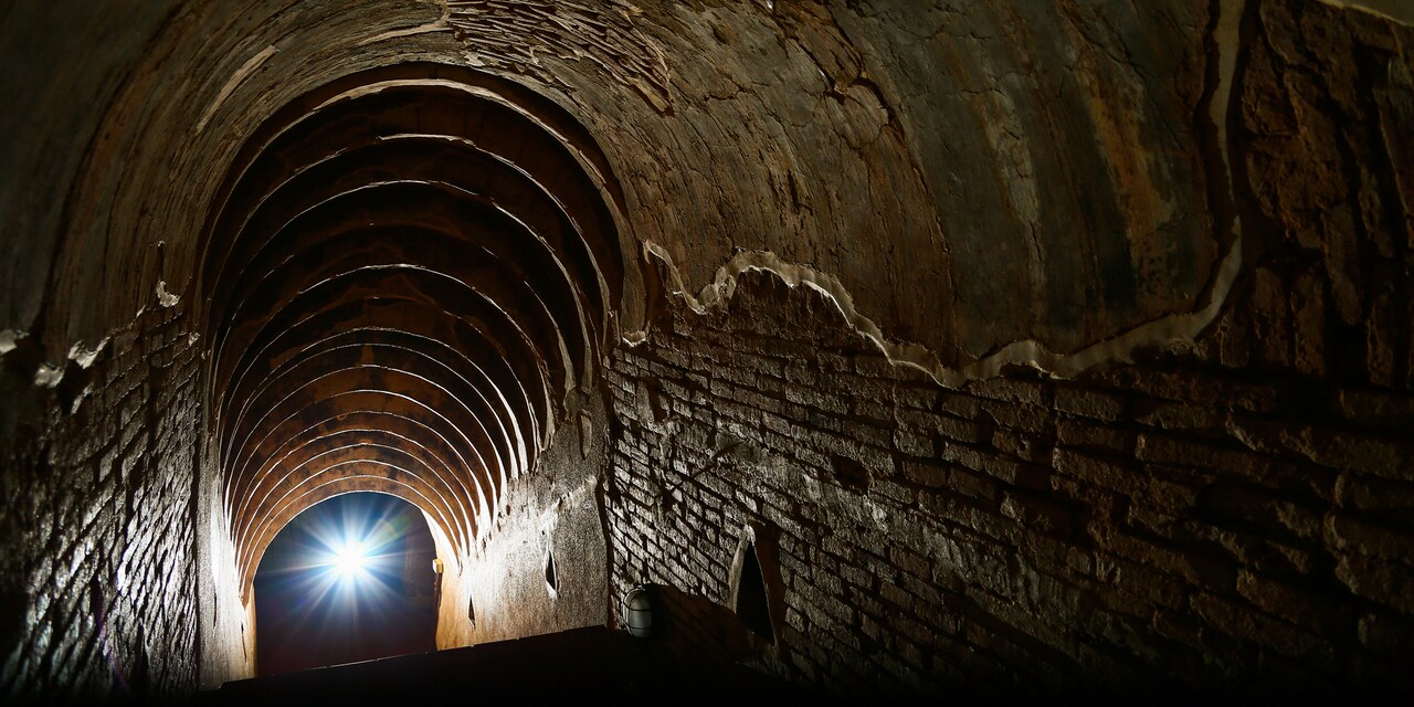 A dark, brick-walled tunnel with an arched ceiling leads downward towards a bright light at the Cú Chi Tunnels