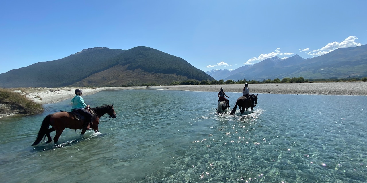 Three people ride horses through a river to a sandy bank
