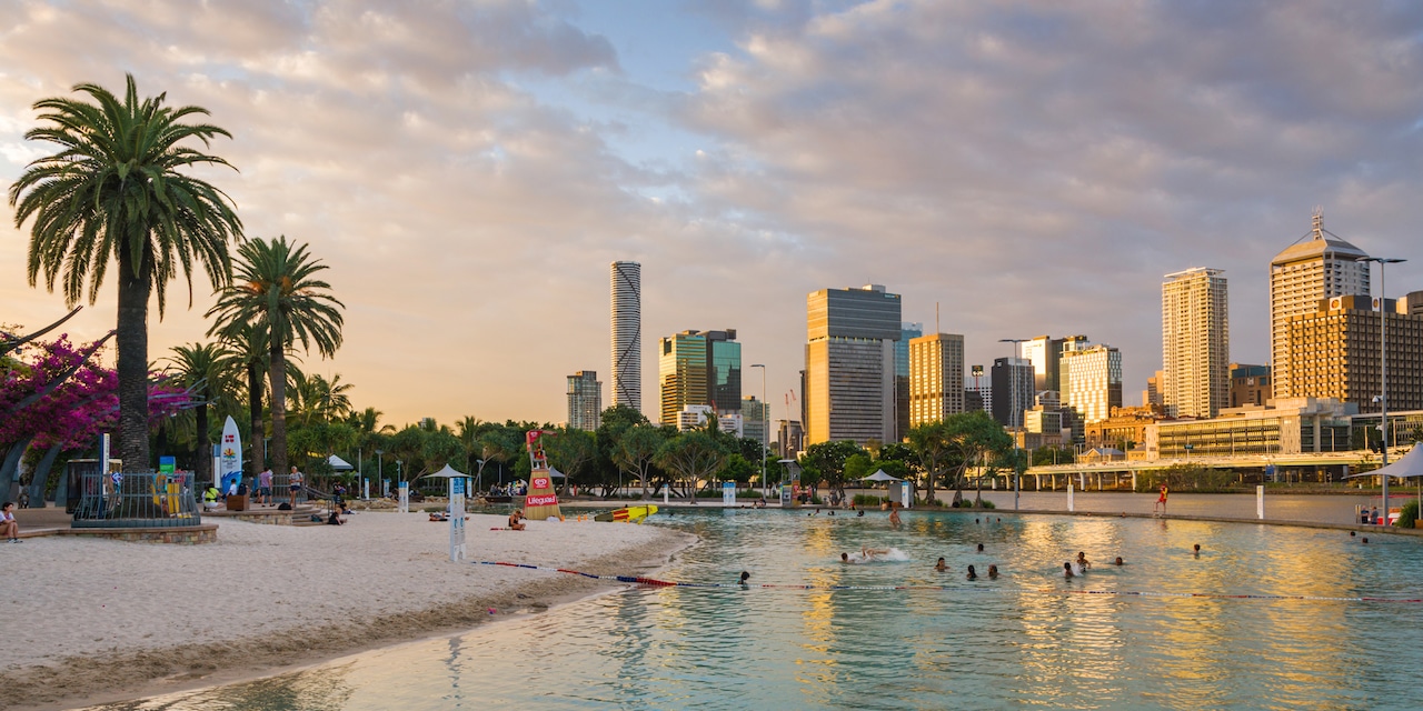 A sandy beach with palm trees on the shores of the Brisbane River near the Brisbane, Australia skyline A sandy beach with palm trees on the shores of the Brisbane River near the Brisbane, Australia skyline