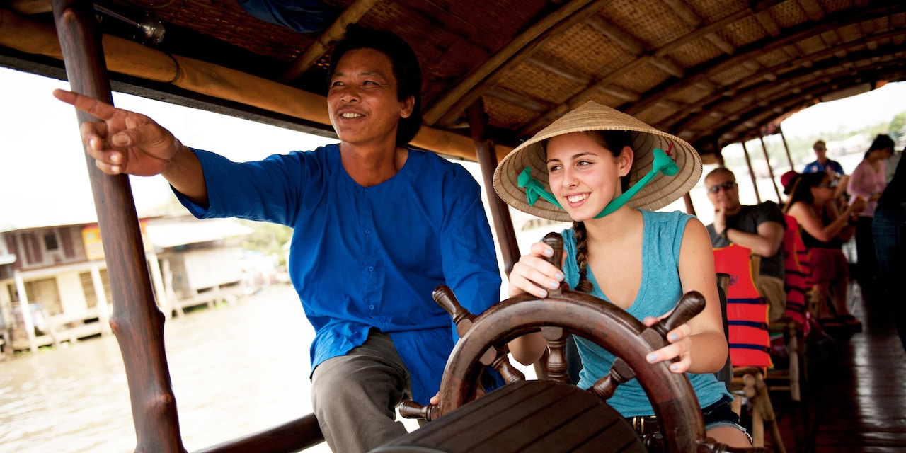 The driver of a roofed touring boat cruising on a river lets a female tourist take the wheel The driver of a roofed touring boat cruising on a river lets a female tourist take the wheel