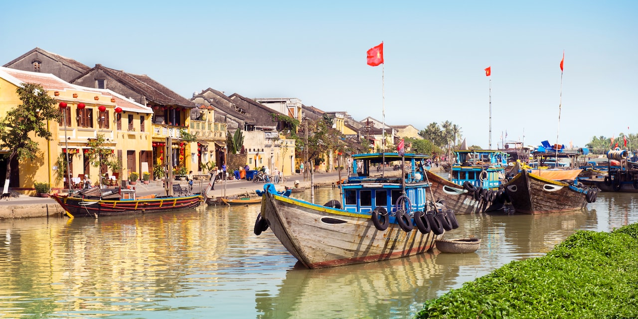 Wooden boats on a river between a grassy riverbank and a building lined riverbank Wooden boats on a river between a grassy riverbank and a building lined riverbank