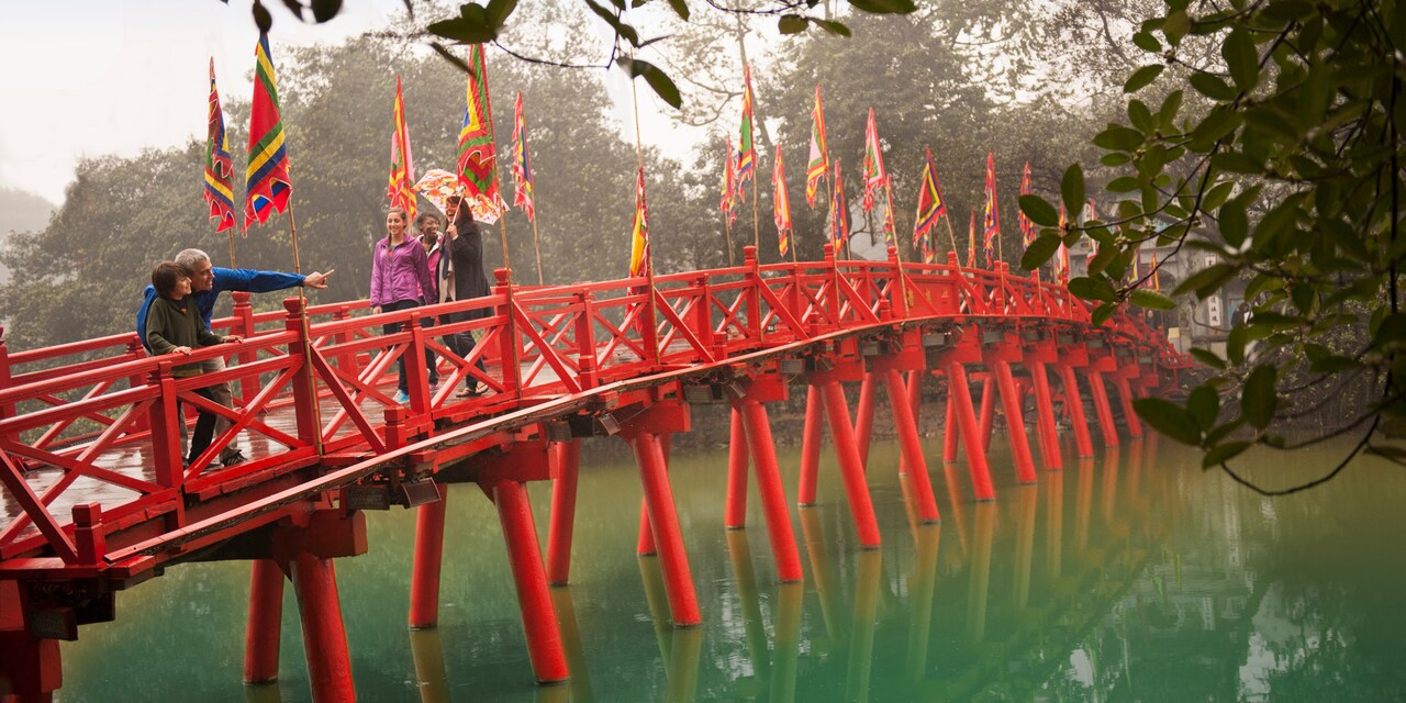 A group of people, including a man pointing something out to a boy, cross over a lake on a bridge that is decorated with flags A group of people, including a man pointing something out to a boy, cross over a lake on a bridge that is decorated with flags