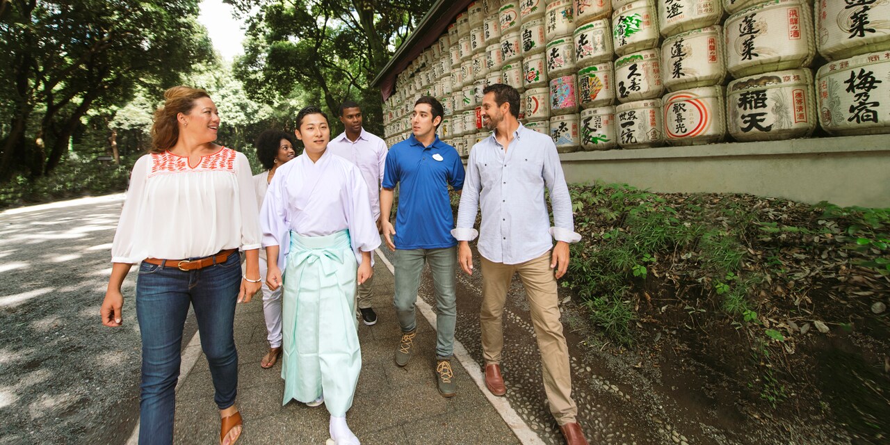 An Adventure Guide leads a group of 5 people on a walk alongside a display of sake barrels decorated with Japanese writing at Meiji Shrine in Tokyo, Japan An Adventure Guide leads a group of 5 people on a walk alongside a display of sake barrels decorated with Japanese writing at Meiji Shrine in Tokyo, Japan