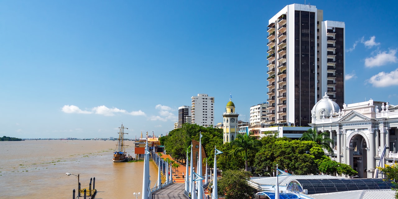 A boat is docked along a boardwalk that stretches between the Guayas River and a building lined street with several high rises and domed buildings A boat is docked along a boardwalk that stretches between the Guayas River and a building lined street with several high rises and domed buildings