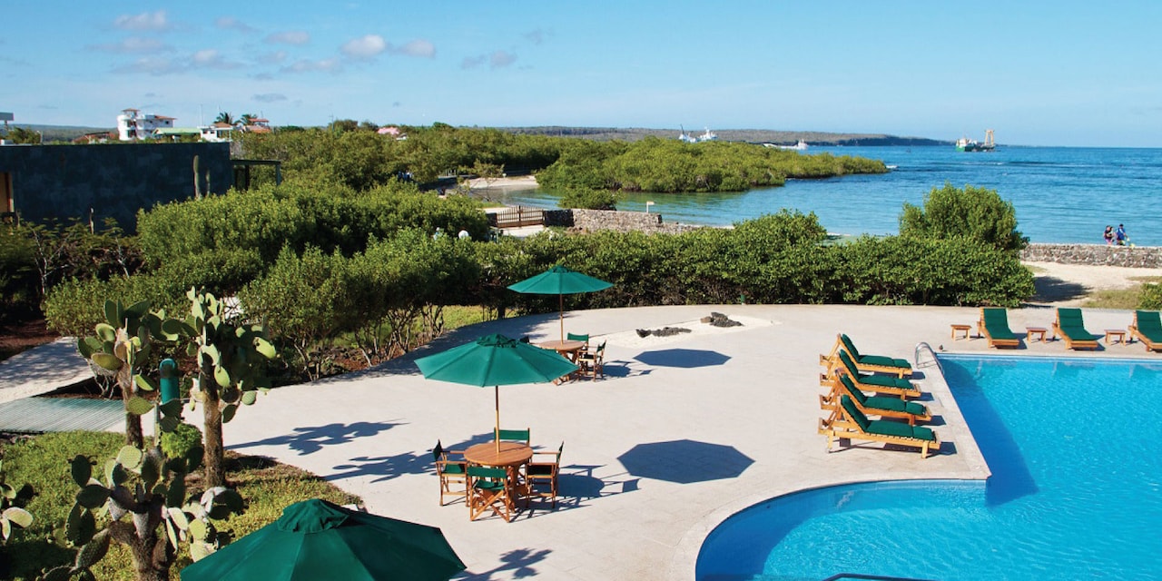 A patio lying between a pool and an ocean, with outdoor recliners and tables with umbrellas A patio lying between a pool and an ocean, with outdoor recliners and tables with umbrellas