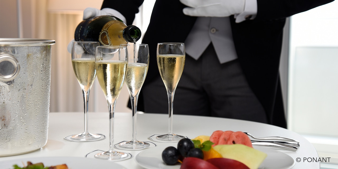 A white gloved server pouring chilled champage into four flutes with a fruit plate on the table