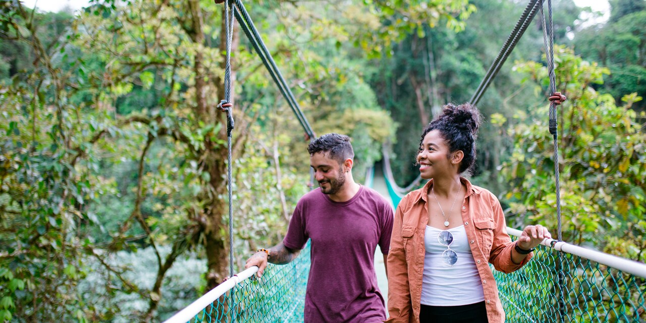 A smiling couple crosses a hanging bridge in a lush nature preserve