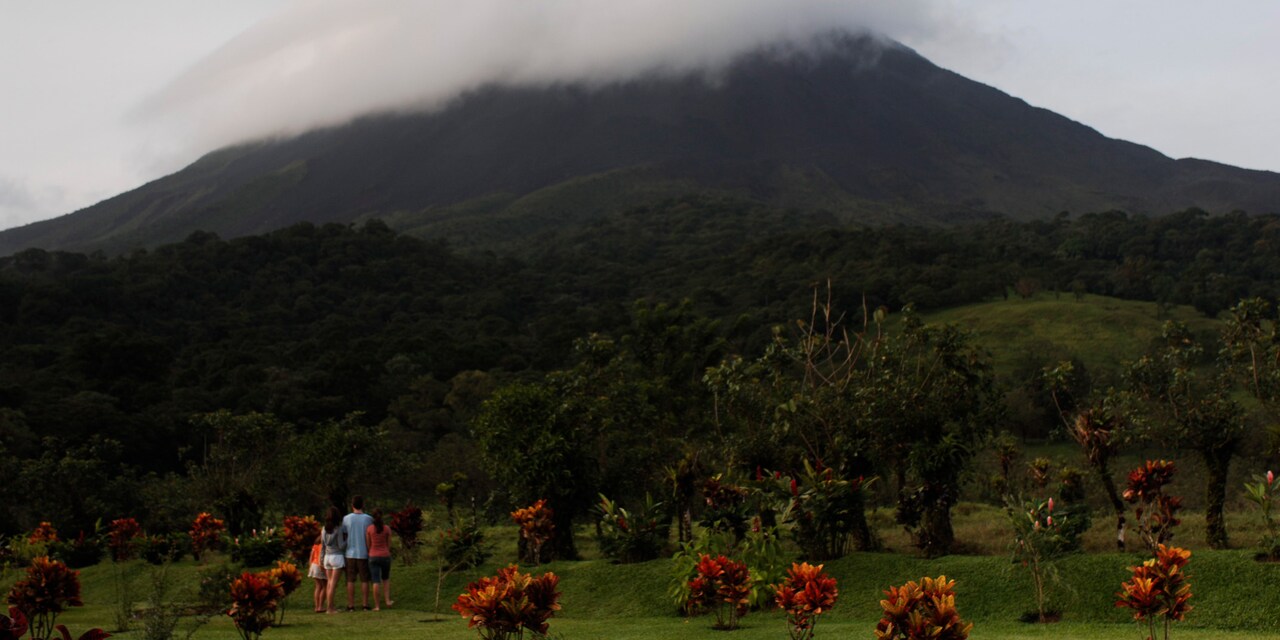 A family of 4 stand in the grass amongst trees looking out at a cloud-covered Arenal Volcano