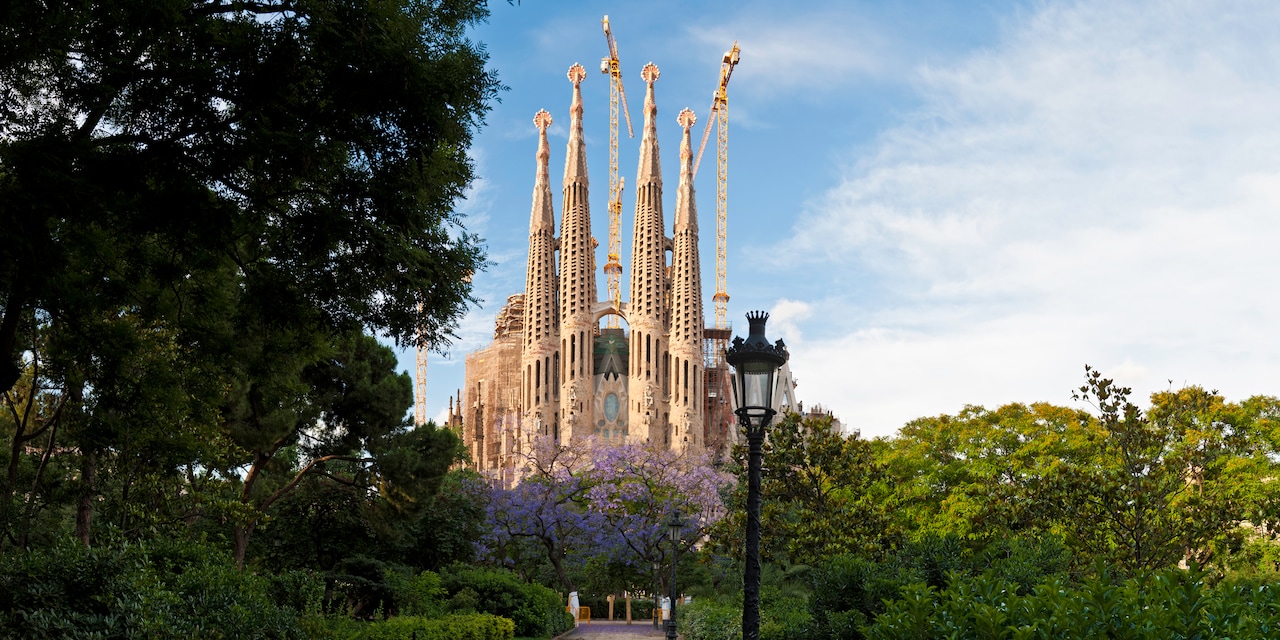 The spires of La Sagrada Familia and some construction cranes stand amid the trees and a lamppost of the surrounding park in Barcelona, Spain The spires of La Sagrada Familia and some construction cranes stand amid the trees and a lamppost of the surrounding park in Barcelona, Spain