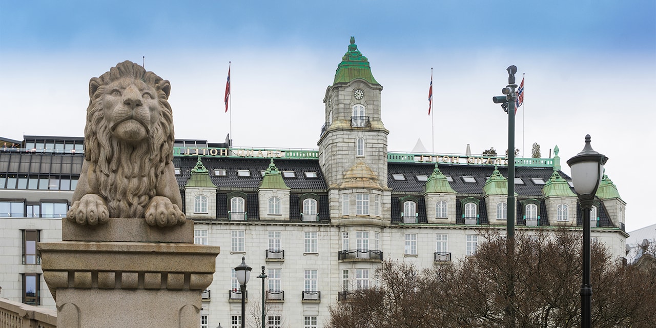 A stone lion perched on a pedestal in front of the exterior façade and clock tower of the Grand Hotel Oslo