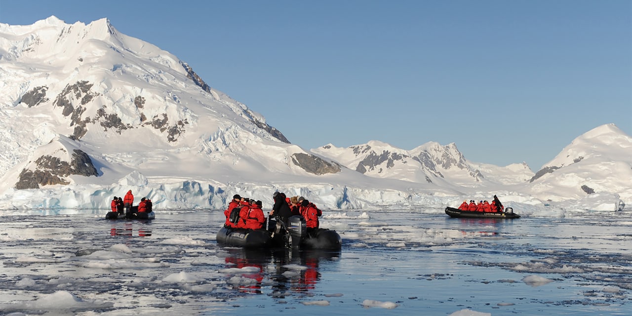 Several inflatable Zodiac boats filled with passengers cruise in the water near a glacier