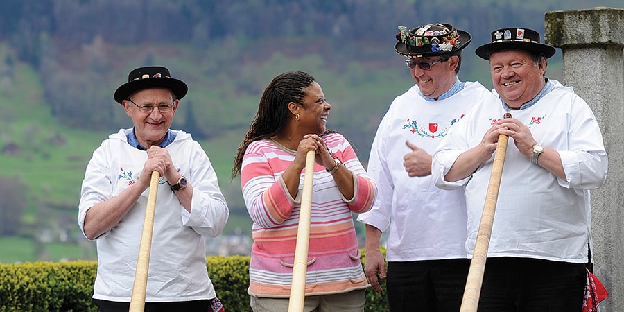 A woman stands with 3 costumed Swiss alpine horn players and laughs as she takes one of their horns A woman stands with 3 costumed Swiss alpine horn players and laughs as she takes one of their horns