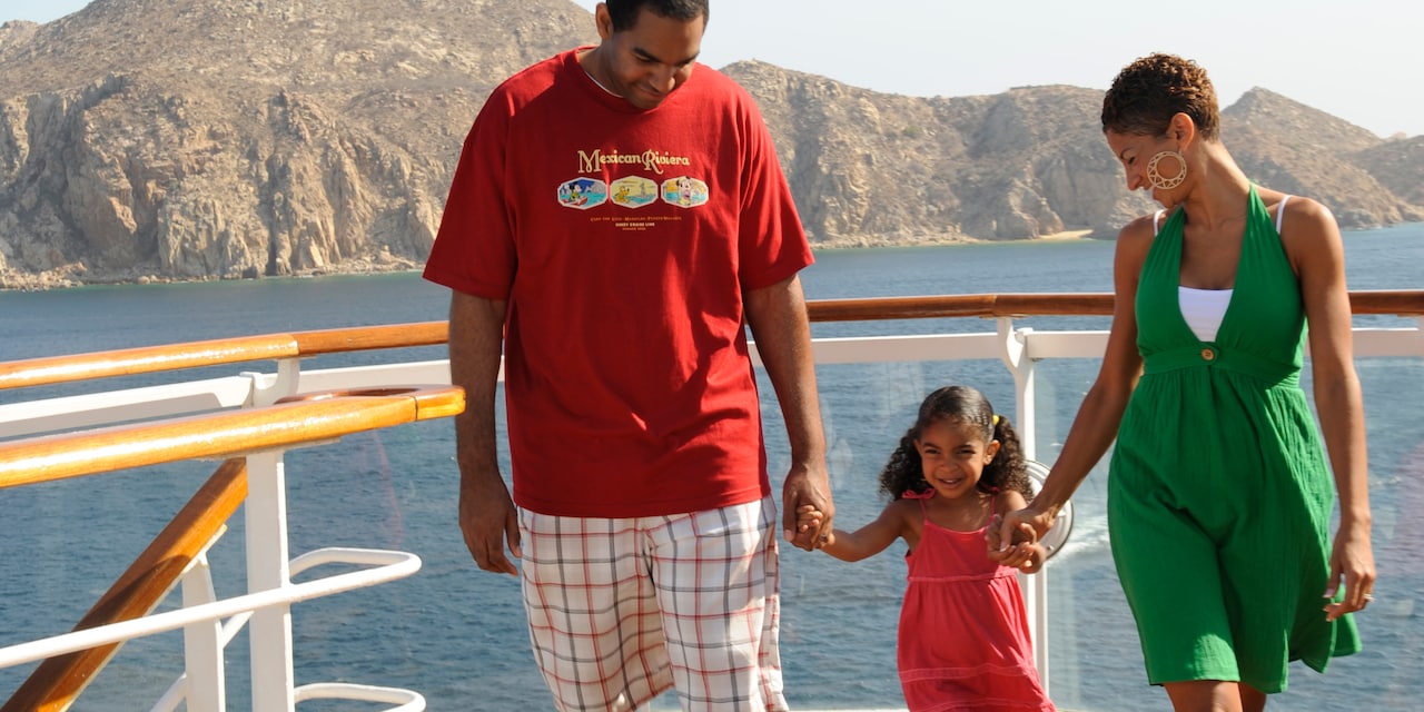 A man and woman hold their young daughter’s hands as they walk on a deck of a ship with a mountain in the background A man and woman hold their young daughter’s hands as they walk on a deck of a ship with a mountain in the background