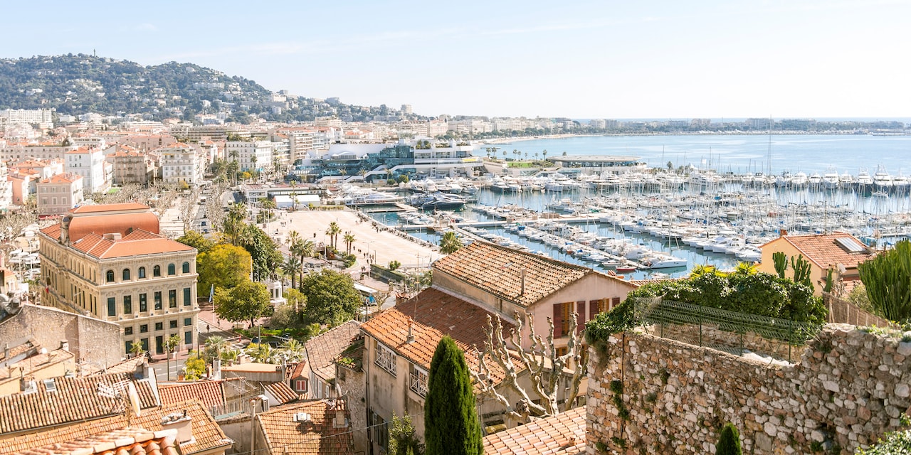 Boats docked in the Cannes Harbor with multi-storied, tile roof buildings hugging the shore Boats docked in the Cannes Harbor with multi-storied, tile roof buildings hugging the shore
