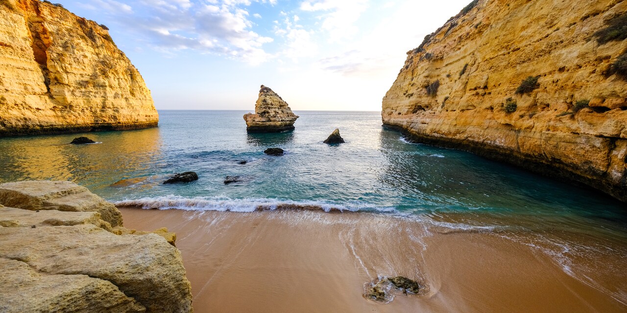 A rock formation in the water between 2 rocky cliffs near a sandy beach