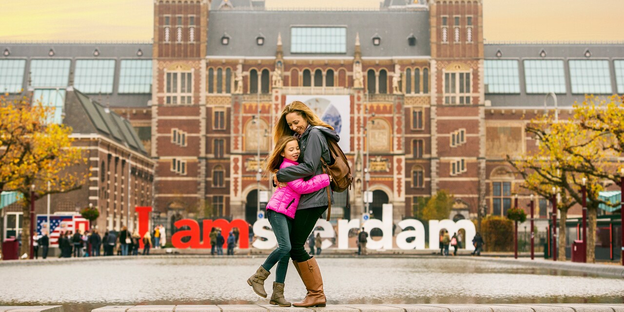 A mother and daughter hug in a square outside a brick building with a sign reading 'I Amsterdam'