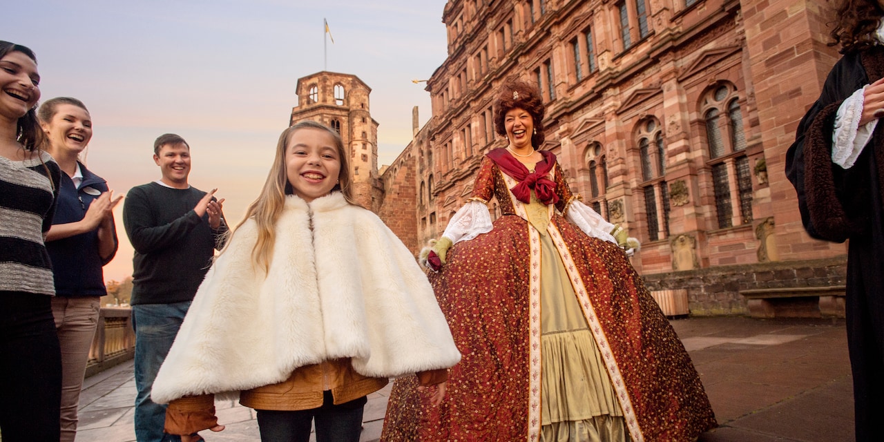 A girl wearing a fur jacket, her family and a woman dressed in a queen's costume outside a castle