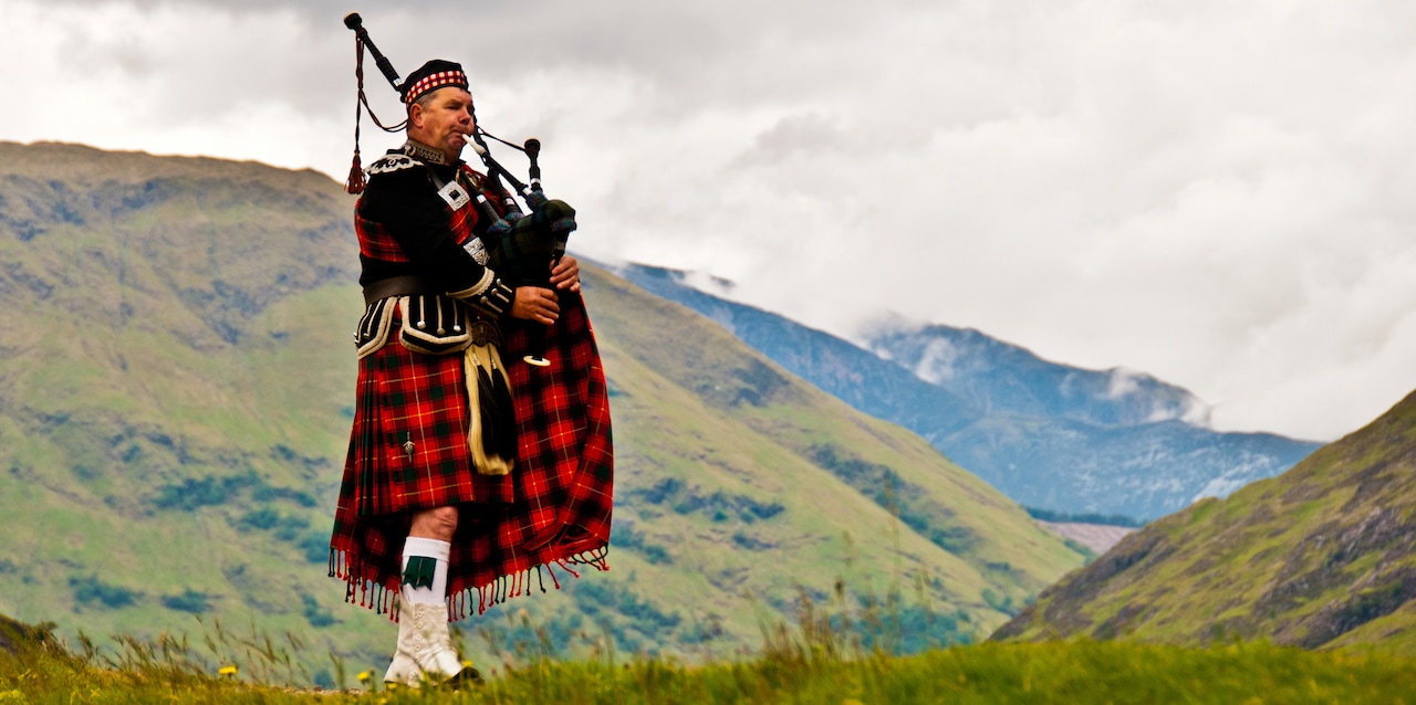 A Scotsman wearing a tartan kilt and traditional Scottish clothing stands in the hills playing a bagpipe