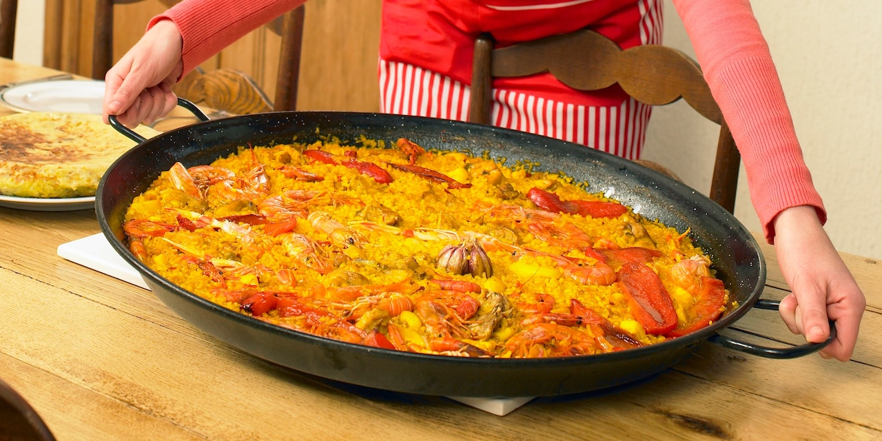 A woman places a large pan of paella on a wooden dinner table