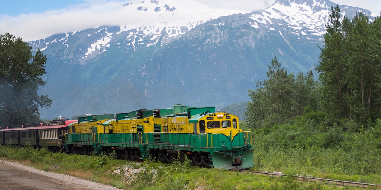 A train traverses the rails over the Alaskan terrain with a mountain range in the background A train traverses the rails over the Alaskan terrain with a mountain range in the background