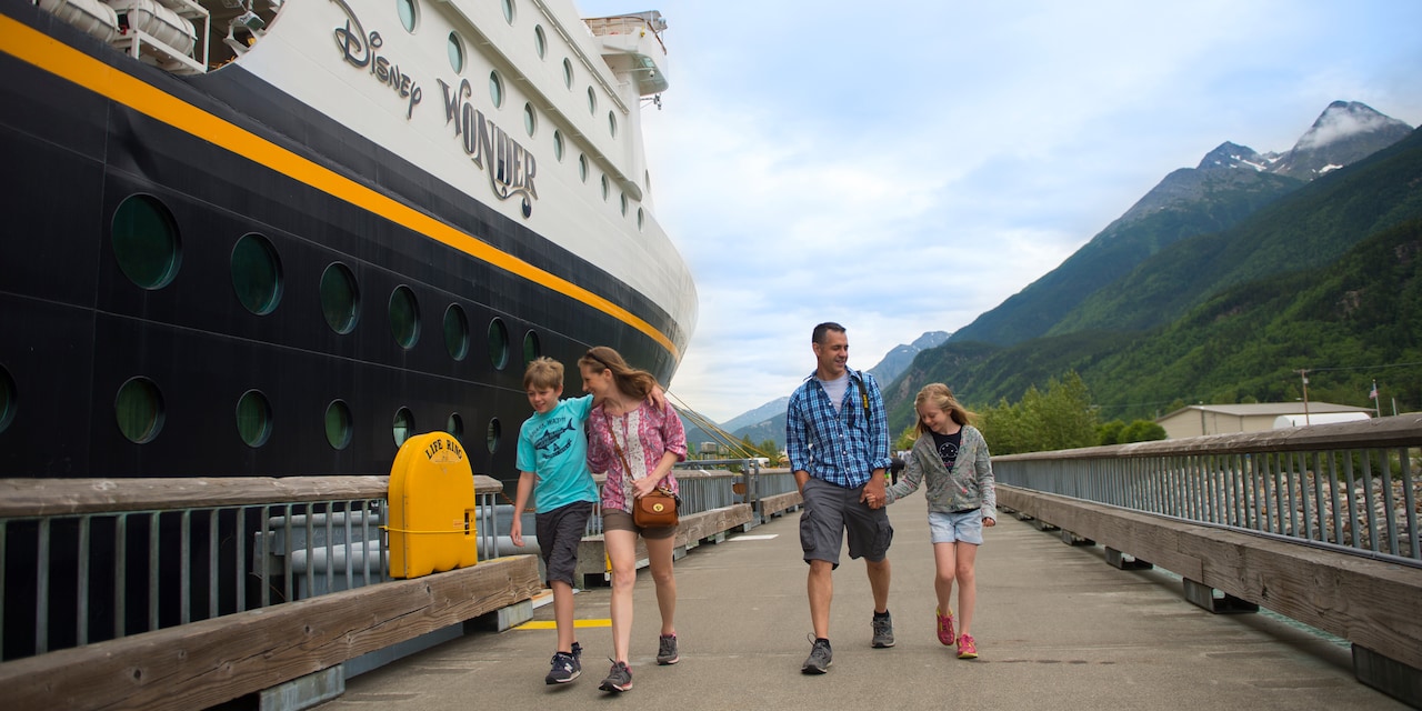 A family of 4 walk along a wood-plank road next to the Disney Wonder cruise ship with snow-capped mountains in the background A family of 4 walk along a wood-plank road next to the Disney Wonder cruise ship with snow-capped mountains in the background