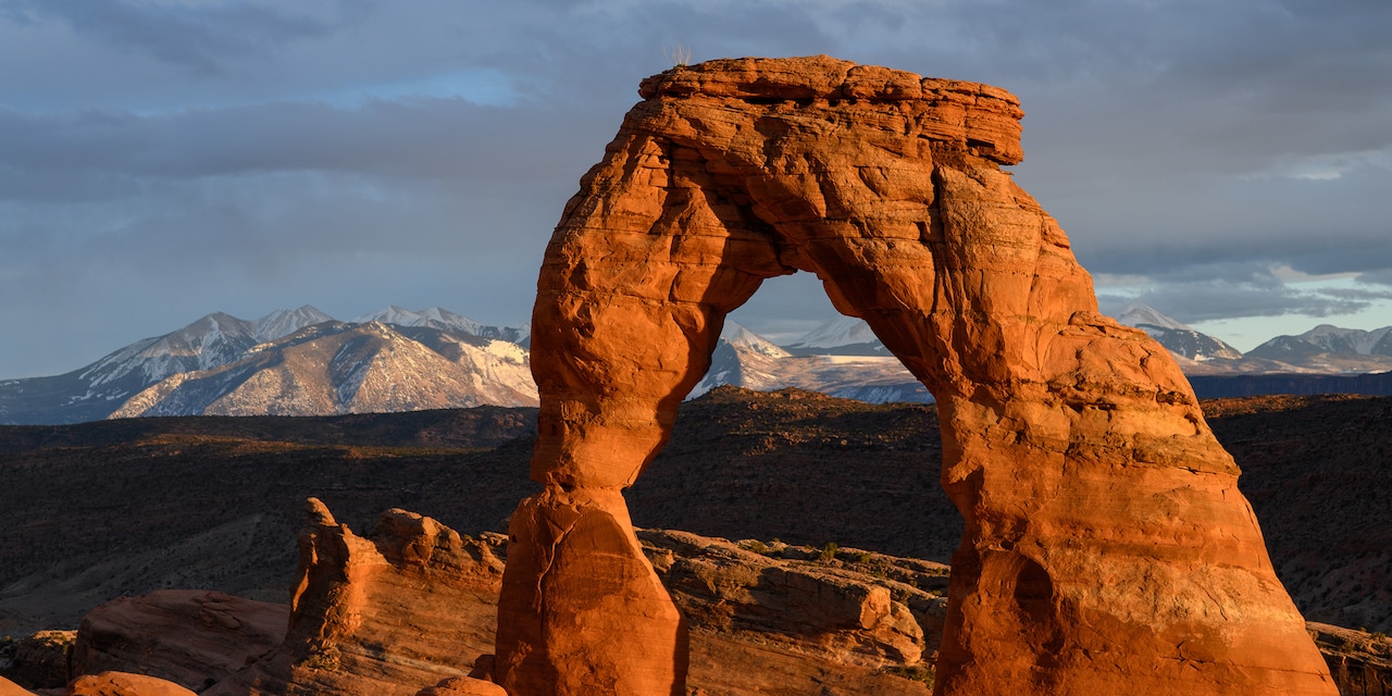 A natural sandstone arch in Arches National Park with snow-capped mountains in the background