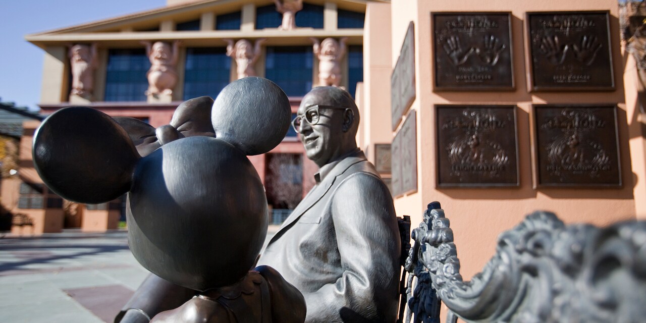 A statue of Roy Disney and Minnie Mouse sitting on a bench near a wall featuring plaques with handprints at The Walt Disney Studios