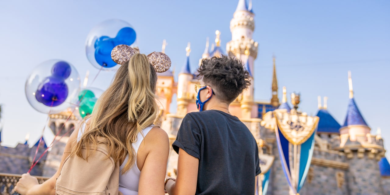 Two women, one wearing a Minnie Mouse ear band and holding Mickey Mouse balloons, stand next to each other facing Sleeping Beauty Castle in Disneyland park.