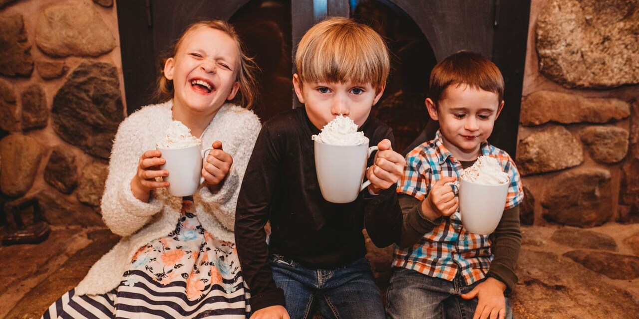 Two boys and a girl sitting down and holding large mugs topped with whipped cream