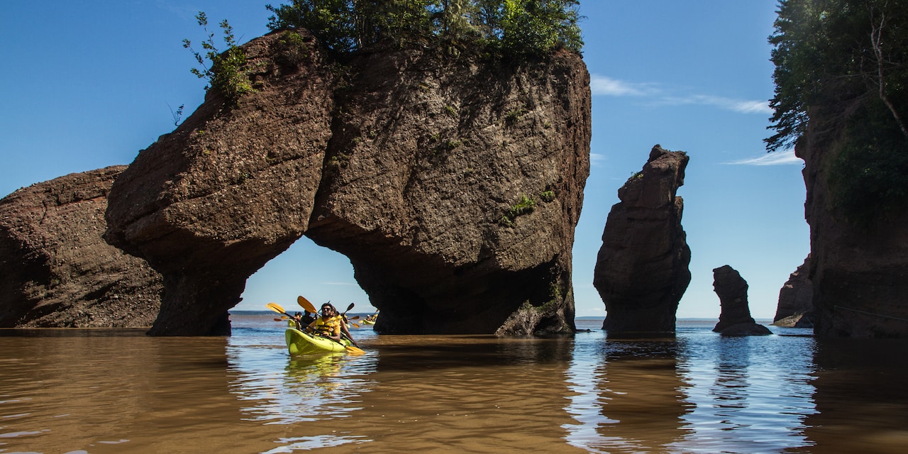 Several kayaks paddle through an arch in a large stone formation near several other rock formations