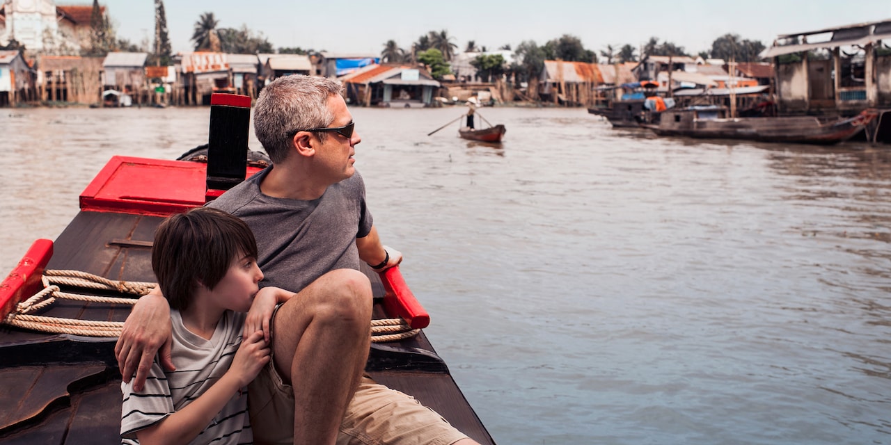 A father and son drift past a Mekong River village while sitting in a canoe