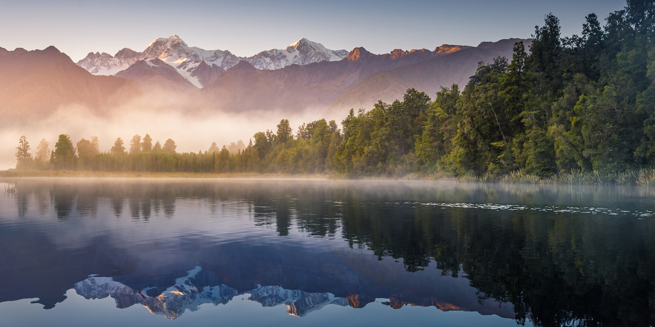 Mist rises from Wanaka Lake with tree covered banks near a mountain range at dawn