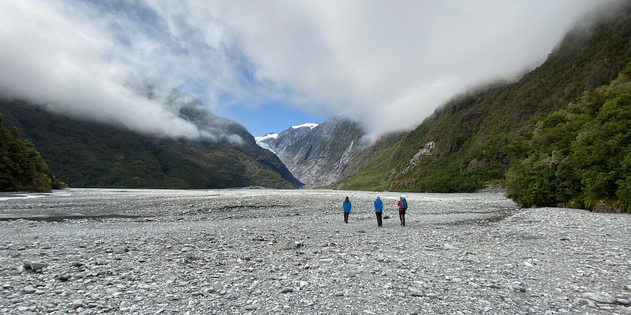 Three people walk on a stone wash towards a glacier between mountains enshrouded by clouds