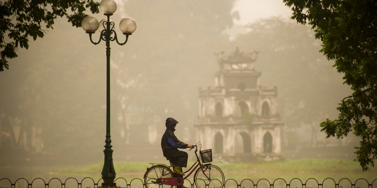 A hooded bicyclist rides past a lamp post on a sidewalk near an old temple in the rain
