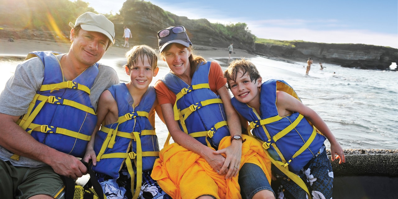 A family of four sitting in front of a beach with lifejackets on