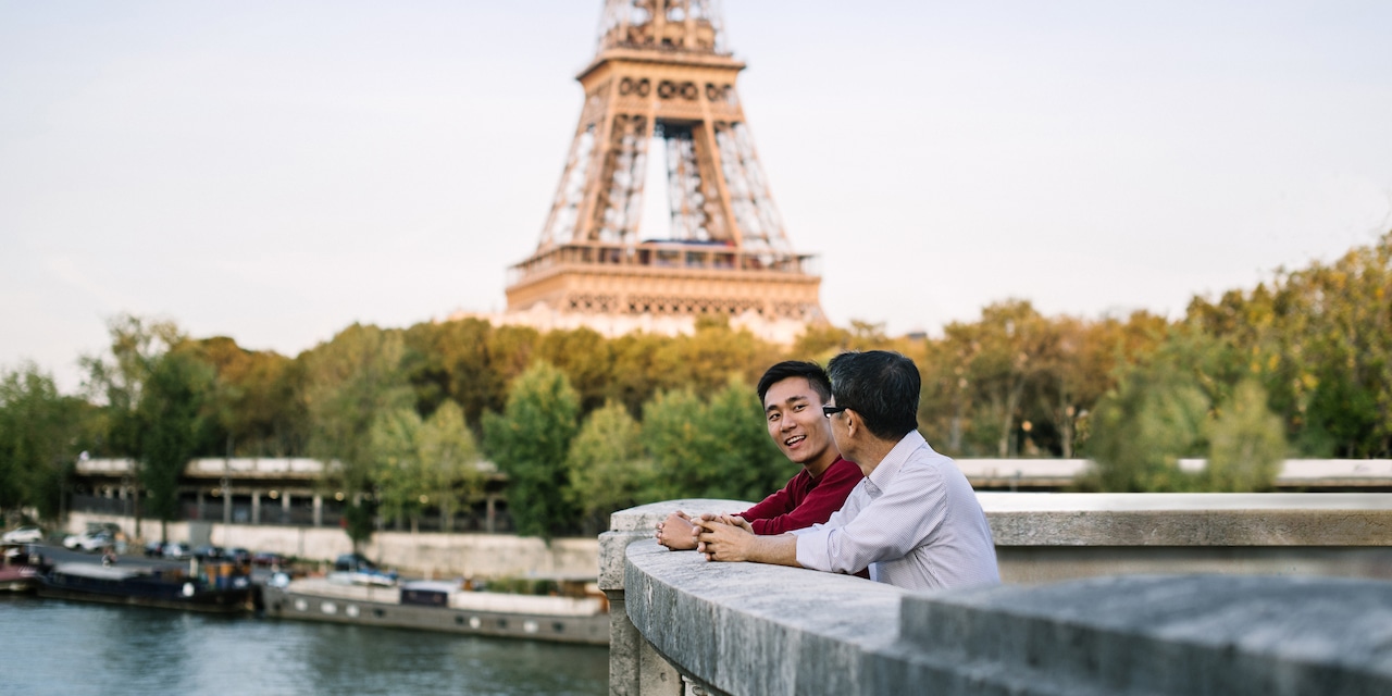 Two men talking with their arms resting on a stone wall that overlooks a river with the Eiffel Tower in the background