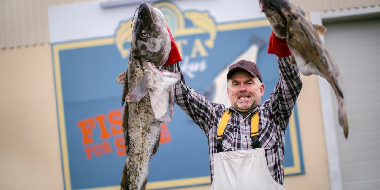 A smiling man wearing a cap and apron holds 2 large fish in front of a sign that reads, “EKTA Fiskus: Fish for Sale”