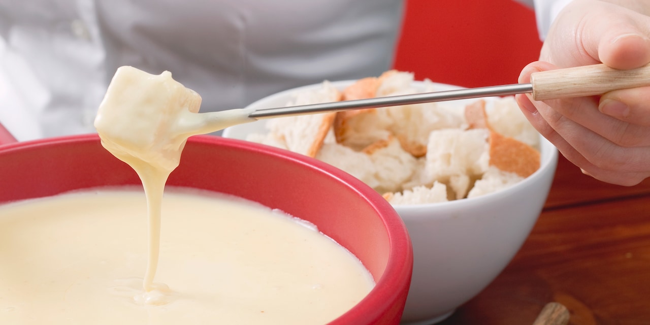A man's hand holds a cube of bread on a long fondue fork that he dips into a bowl of fondue