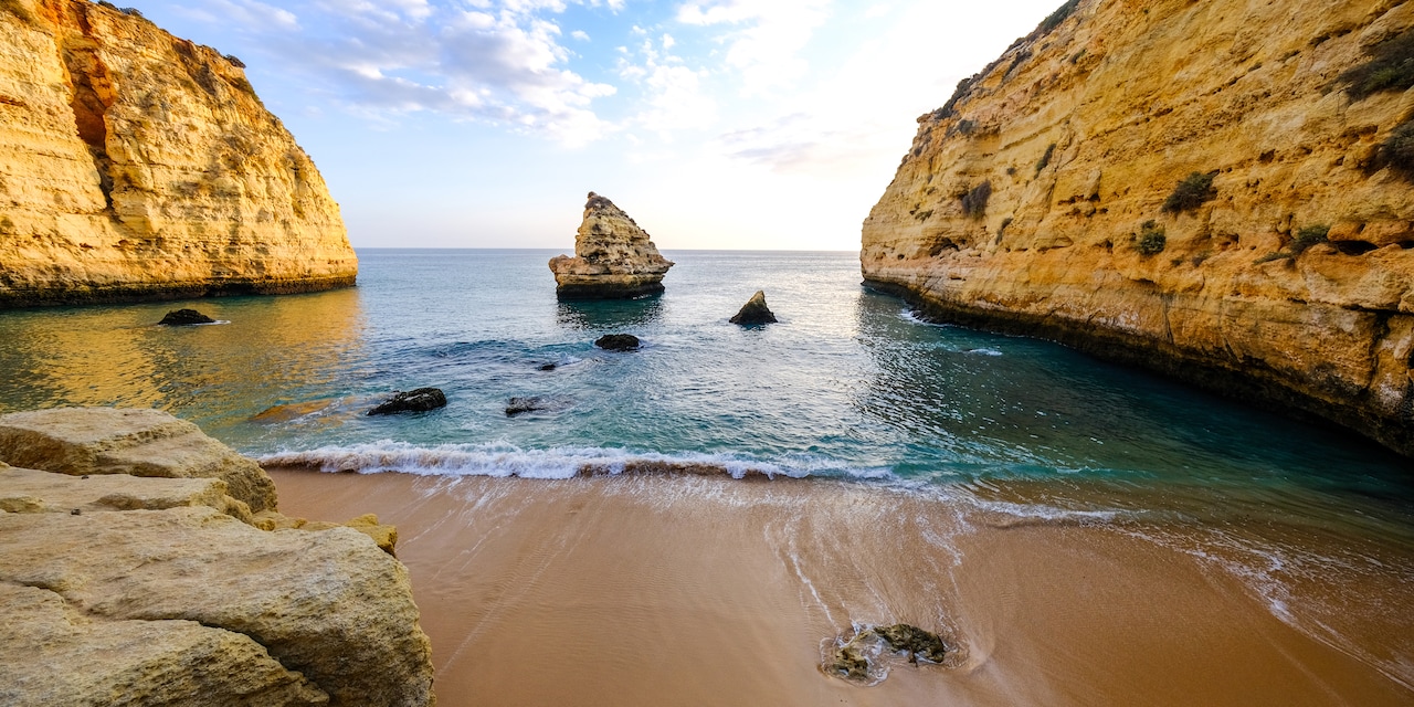 A rock formation in the water between 2 rocky cliffs near a sandy beach