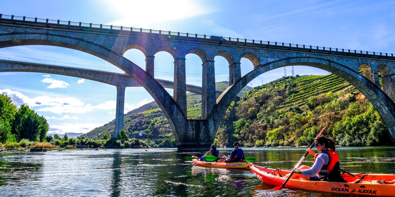 Four people paddle 2 kayaks on a river near an arched bridge