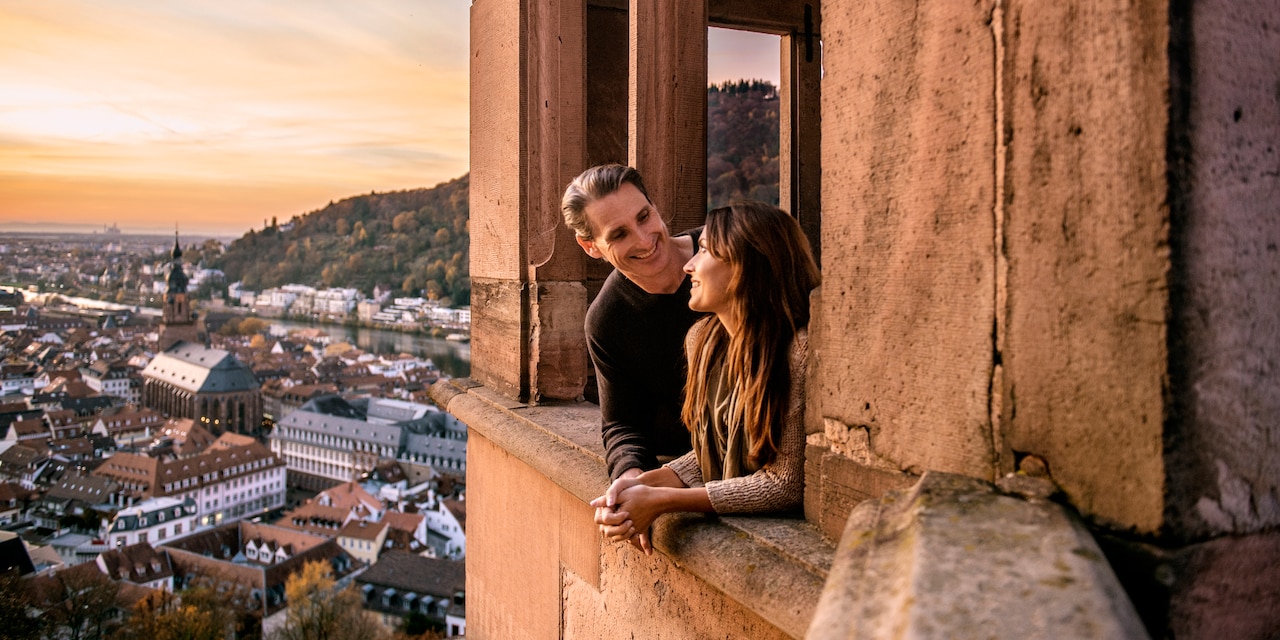 A man and woman smile at each other as they lean out a window overlooking an old European town 
