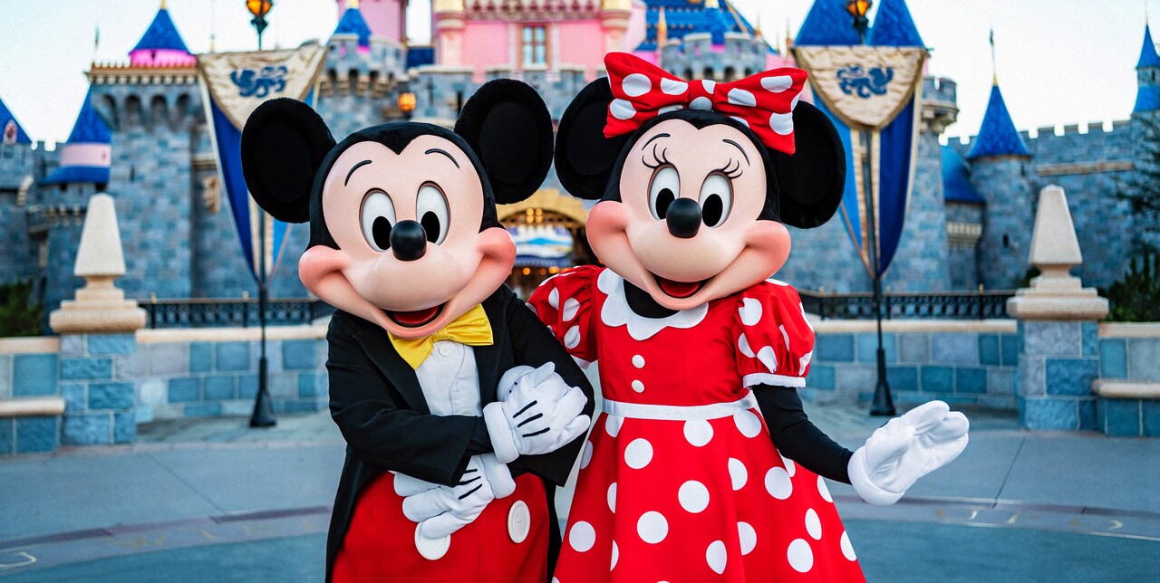 Mickey Mouse and Minnie Mouse stand arm in arm in front of Sleeping Beauty Castle at Disneyland Park
