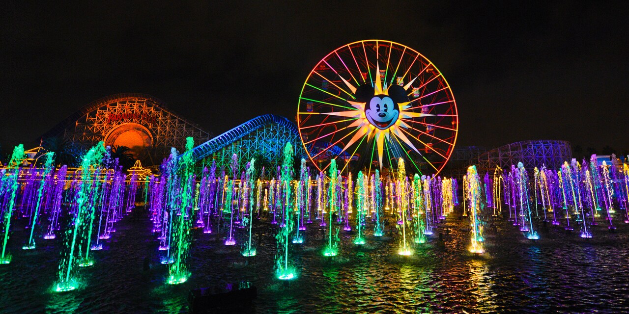 Fountains of water spray into the nighttime air with a large wheel with Micky Mouse’s face in the background
