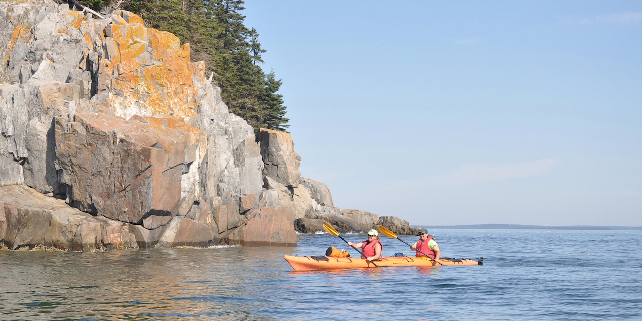 Two people paddling a two-person kayak near a rocky outcropping in a lake